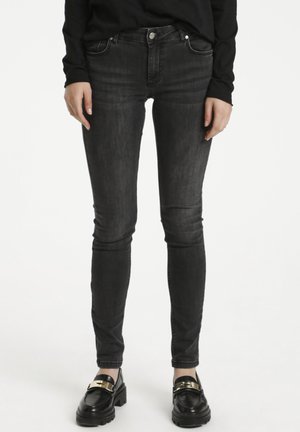 Osoba w dopasowanych, przetartych czarnych dżinsach i czarnych mokasynach z złotą klamrą, stojąca na białym tle, z rękami swobodnie opuszczonymi wzdłuż ciała.