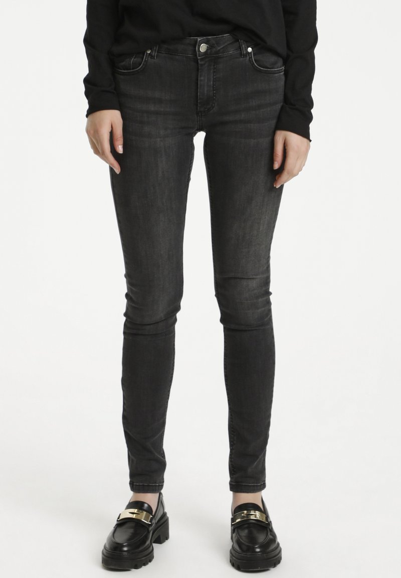 Personne portant un jean noir délavé ajusté et des mocassins noirs avec une boucle dorée, debout contre un fond blanc avec les mains détendues le long du corps.