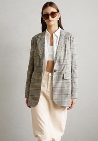 Grauer Karoblazer mit Schulterpolstern, einreihiger Knopfverschluss, mit Revers, kombiniert mit einem weißen Cropped-Top und cremefarbenen Hosen.