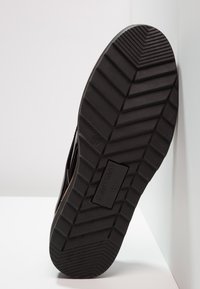 Semelle en caoutchouc noir avec un motif texturé, présentant de profondes rainures et une étiquette de marque rectangulaire. Légèrement inclinée sur un fond blanc.