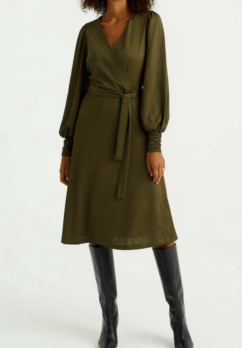 Olivgrön omlottklänning i texturerat tyg, långa puffärmar och knytmidja. Bärs med svarta knähöga stövlar.