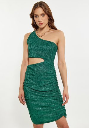 Frau trägt ein glänzendes grünes One-Shoulder-Kleid mit Ausschnitt an der Taille und posiert vor einem schlichten weißen Hintergrund.