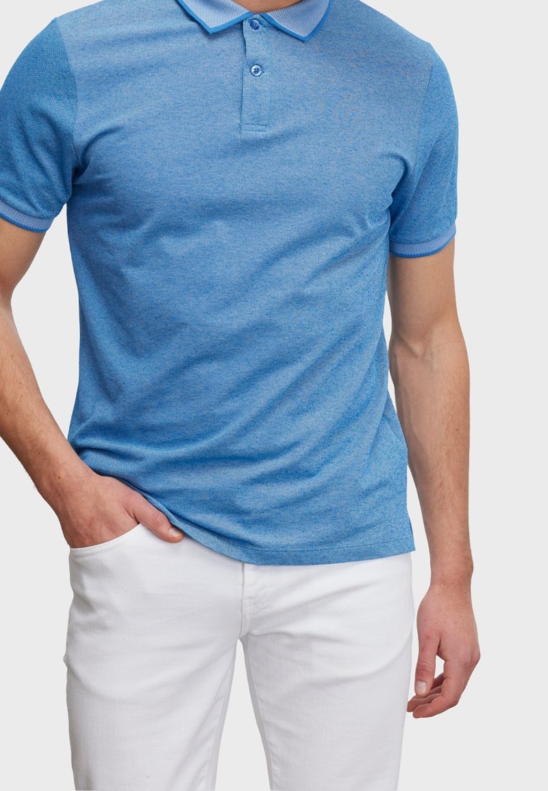 Camiseta polo azul de manga corta, tela texturizada, tapeta con dos botones y cuello y puños en contraste. Combinada con pantalones blancos.