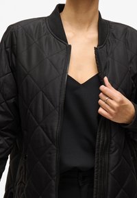 Černá prošívaná bomber bunda se zapínáním na přední zip, žebrovaným límcem a dvěma bočními kapsami, nošená přes černý top. Hladká textura látky.