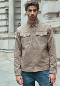 Jachetă din suede bej cu închidere cu butoane, două buzunare pe piept, butoane metalice argintii și guler. Purtați peste blugi din denim albastru.