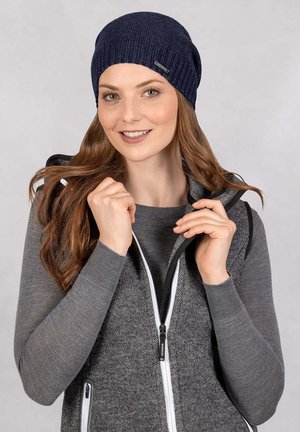 Marineblauwe ribgebreide muts met een gladde textuur, gecombineerd met een grijze sleeveless jas met contrasterende witte ritsen en een gestructureerde stof.