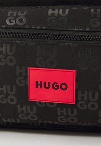 Fekete anyagú táska, domború "HUGO" mintával, piros téglalap alakú logóval fekete felirattal és cipzár részlettel a tetején.