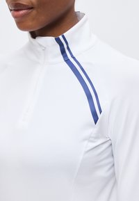 Κοντινό πλάνο ενός ατόμου που φοράει λευκό τοπ με φερμουάρ στο τέταρτο μέρος, ψηλό κολάρο και δύο παράλληλες μπλε ρίγες στον ώμο.