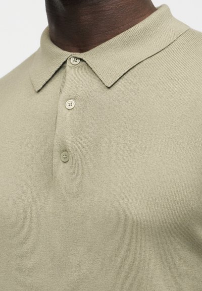 Gros plan sur une personne à la peau foncée portant un polo en maille beige avec une patte de boutonnage à trois boutons et un col pointu.
