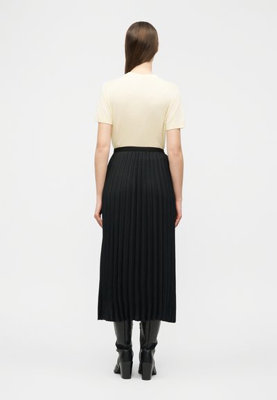 Γυναίκα που στέκεται με την πλάτη στην κάμερα, φορώντας εκρού τοπ με κοντά μανίκια, μαύρη πλισέ μίντι φούστα και μαύρες μπότες με τακούνι, σε λευκό φόντο.