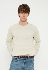 Fiatal férfi göndör hajjal, krémszínű BOSS pulóvert, kék farmert és barna fonott övet viselve, sima fehér háttér előtt áll.