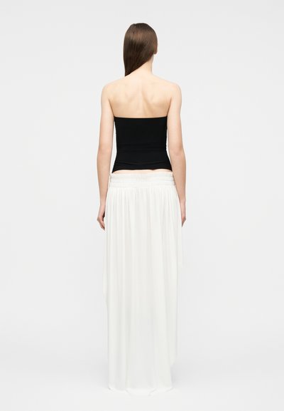 Γυναίκα που στέκεται γυρισμένη με την πλάτη, φοράει μαύρο τοπ χωρίς τιράντες και μακριά άσπρη ροή φούστα, μπροστά σε απλό φόντο.