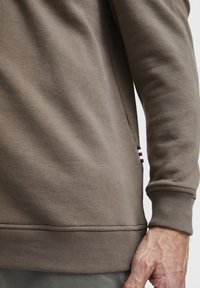 Sudadera marrón con puños y dobladillo acanalados, que presenta un acento en la costura lateral con franjas azul, blanca y roja. Tejido suave y texturizado.