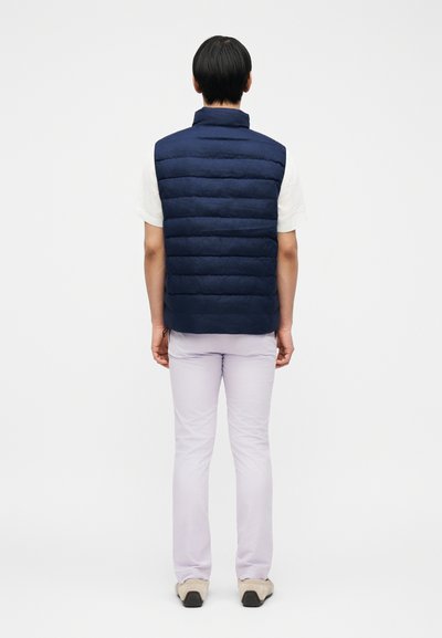 Άτομο που στέκεται πλάτη, φορώντας μπλε καπιτονέ γιλέκο πάνω από λευκό κοντομάνικο πουκάμισο, ανοιχτόχρωμο παντελόνι και μπεζ παπούτσια.