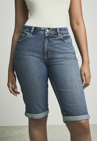 Pantalones cortos de mezclilla en azul oscuro con cintura alta, que presentan dobladillos enrollados y un ligero desgaste. Diseño de cinco bolsillos con herrajes plateados.