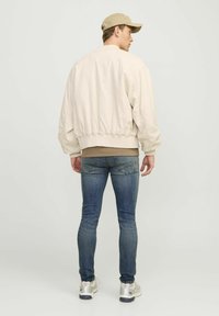 Chaqueta bomber beige con puños y dobladillo elásticos, combinada con skinny jeans de color azul oscuro y zapatillas beige, vista desde atrás.