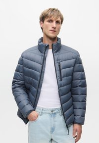 Párnázott dzseki szürke-kék színben, magas gallérral, cipzáras elülső résszel és mellzsebbekkel. Az anyag sima, steppelt részletekkel.