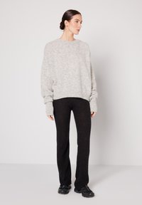 Naine kannab lahtist helehalli kampsunit, musti kitsaid pükse ja musti spordijalatseid, seistes ühevärvilise valge tausta vastas.