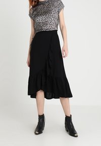 Sort indpakningsskørt med bølget kant, parret med en grå leopardprint top. Modellen har sorte ankelstøvler med gyldne detaljer på.