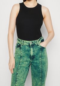 Žena v bezramenném černém topu a vysokých acid-wash zelených džínách, s jednou rukou v kapse, na jednobarevném pozadí.