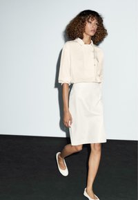 Woman with curly hair walking in a cream button-up blouse and matching knee-length skirt with white flat shoes on a black floor.