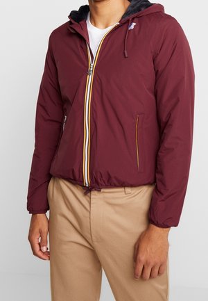 Homme portant une veste à capuche bordeaux zippée avec des bordures blanches et jaunes, un pantalon beige et une chemise blanche en dessous.