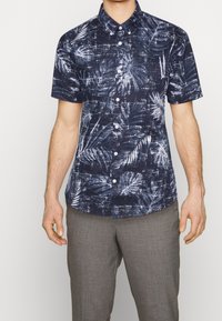 Chemise à manches courtes, bleu marine, boutonnière avec un motif de feuilles de palmier blanches, tissu texturé et col classique. Associée à un pantalon gris.