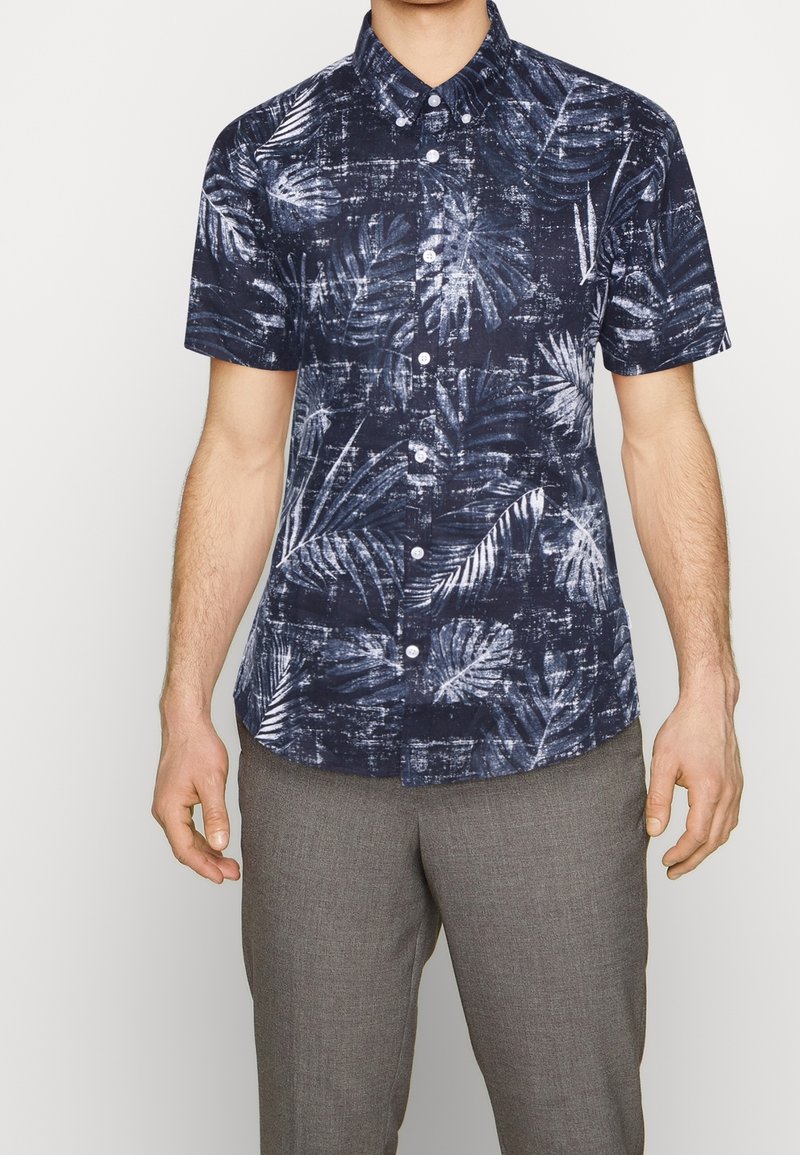 Camicetta a maniche corte, con bottoni, di colore blu navy, decorata con un motivo di foglie di palma bianche, in tessuto strutturato e con colletto classico. Abbinata a pantaloni grigi.