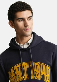 Junger Mann mit kurzen lockigen Haaren, der einen navyblauen Kapuzenpulli mit gelber Aufschrift "GANT 1949" und ein weißes Hemd darunter trägt.