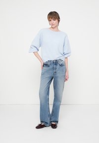 Jasnoniebieski sweter z bufkami, w zestawie z jasnoniebieskimi spodniami o wysokim stanie. Modelka nosi błyszczące burgundowe baleriny. Minimalistyczne tło.