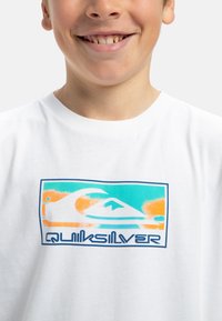 Vitt bomullströja med en flerfärgad grafisk design som har vågor och bergsmotiv. Märkesnamnet "Quiksilver" syns.