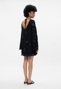 Melnā kleita ar garām piedurknēm, ziedu izšuvumiem, dziļu V formas aizmuguri un caurspīdīgu pārklājumu. Apvienota ar zemu papēžu melnajām sandalēm.