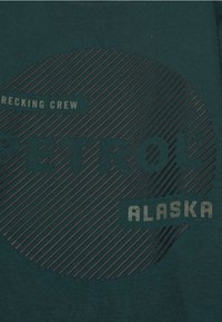 Sötétzöld póló textúrált grafikai mintával. Tartalmazza a "PETROL" és "ALASKA" feliratokat eltérő betűtípusokban, valamint csíkos háttérmintát.
