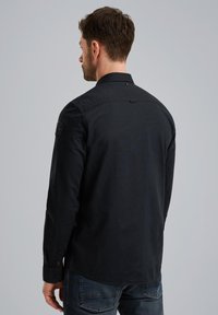 Camisa negra de manga larga con cuello abotonado, que presenta un parche en el hombro izquierdo y un dobladillo redondeado, hecha de una tela texturizada.
