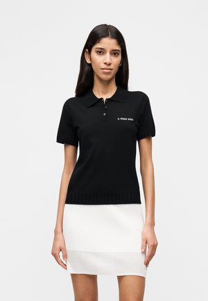Femme portant un polo noir à manches courtes avec des boutons et un texte blanc, associé à une mini-jupe blanche côtelée, posant devant un fond uni.