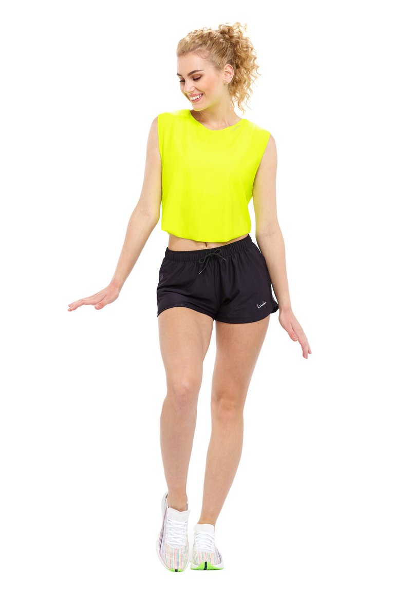 Top cortado en amarillo neón, pantalones cortos deportivos negros con cordón, y zapatillas blancas con un patrón y acentos verdes, llevadas por una persona en movimiento.
