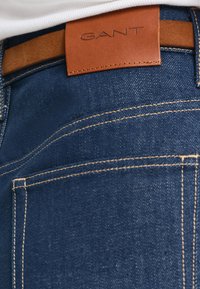 Jeans in denim blu scuro con cuciture visibili e un'etichetta in pelle marrone. L'etichetta presenta il nome del marchio "GANT" in rilievo.