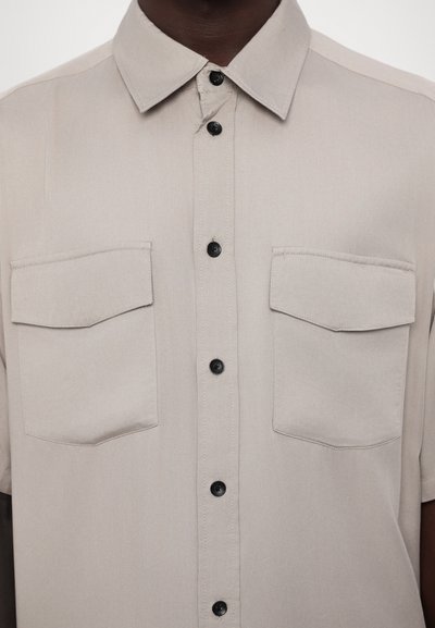Chemise à manches courtes en tissu beige clair, dotée d'une fermeture à boutons sur le devant, de deux poches poitrine et de boutons noirs pour le contraste.