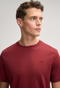 Bordeauxfarbendes Baumwoll-T-Shirt mit Rundhalsausschnitt, kurzen Ärmeln und einem kleinen gestickten Logo auf der Brust. Glatte Textur, minimalistisches Design.