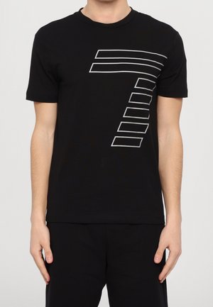 Personne portant un t-shirt noir à manches courtes avec un design de numéro 7 en rayures géométriques blanches sur la poitrine, associé à un pantalon noir.