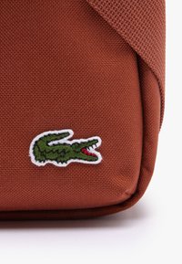 Texturerad, rostfärgad väska i vävt material med en grön broderad krokodil-logotyp med röda detaljer på en vit kant.