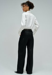 Camisa blanca de manga larga con puños abotonados, combinada con pantalones negros de pierna ancha que presentan un detalle de cadena dorada en la cintura. Texturas suaves.