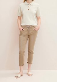 Polo crème à manches courtes avec trois boutons marron, associé à un pantalon tapered beige clair et des sandales beige à enfiler.