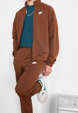 Personne portant un survêtement Nike marron avec un t-shirt bleu sarcelle, des baskets Nike blanches et vertes, s'appuyant contre un mur blanc avec une jambe levée.