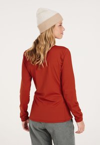 Langarmshirt in roestkleur, gemaakt van gladde stof. Gelaagd met een beige gebreide beanie met een omgevouwen rand. Zichtbaar detail van de naad aan de achterkant.