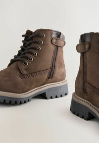Botas de tobillo de ante marrón con cordones en la parte delantera y cremalleras en los laterales. Presentan una suela robusta con bandas pronunciadas y acentos de costura en contraste.
