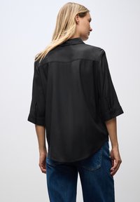 Czarna satynowa bluzka z kołnierzykiem, trzyczwartową długością rękawów i lekko zaokrąglonym hemem. Tkanina ma gładką, błyszczącą teksturę.