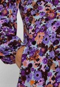 Material cu un model floral în nuanțe de violet, albastru și portocaliu. Mâneci lungi, bufante, cu manșete strânse și o croială lejeră.