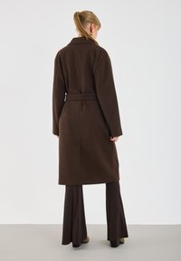 Donna con coda di cavallo che indossa un lungo cappotto marrone scuro con cintura, pantaloni a campana abbinati e orecchini a cerchio dorati, in piedi su uno sfondo neutro.