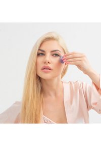 Fioletowe narzędzie do pielęgnacji twarzy z silikonu trzymane przy policzku, podczas gdy osoba ma na sobie jasnoróżowy satynowy szlafrok z koronkowymi akcentami. Gładkie włosy i neutralny makijaż.